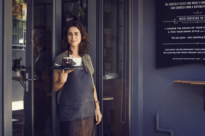 Portrait of confident female owner holding tray with coffee cup outside cafe