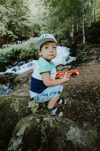 Side view of boy holding toy car while sitting on rock in forest