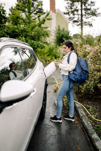 Side view of woman using smart phone while charging electric car