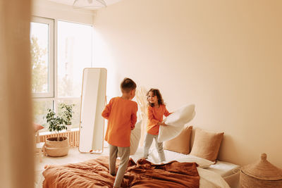 Funny happy kids brother and sister in pajamas playing fighting with pillows in cozy bedroom at home