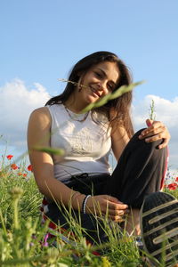 Beautiful young woman sitting on field against sky