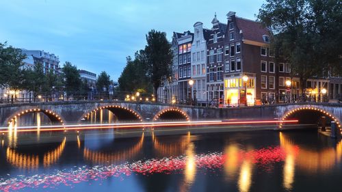 Illuminated bridge over river by buildings against sky at night