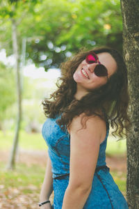 Portrait of smiling young woman in sunglasses standing against tree