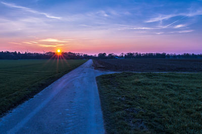 Road by agricultural field against sky during sunset