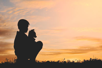 Side view of silhouette boy with dog sitting on field at sunset