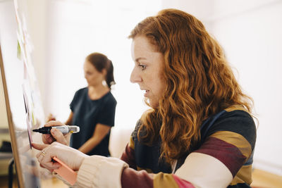 Side view of confident redhead businesswoman writing on whiteboard while female colleague working at creative office