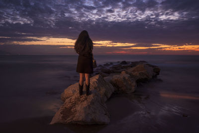 Woman standing on rock at beach against sky during sunset