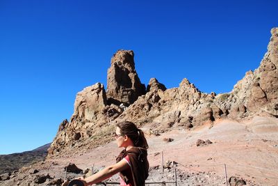 Low angle view of woman sitting on cliff against clear blue sky