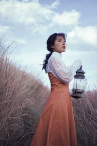 Woman holding old lantern while standing by plants against sky