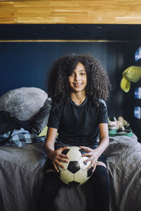 Portrait of curly hair girl with soccer ball sitting on bed at home