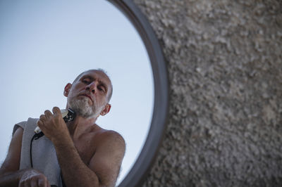 Shirtless adult man shaving himself with shaver in mirror