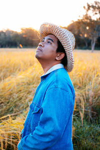 Side view of thoughtful young man wearing hat while standing on field during sunset
