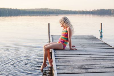  blonde girl child sitting on pier by lake.  laughing kid in swimsuit splashing with legs in water. 