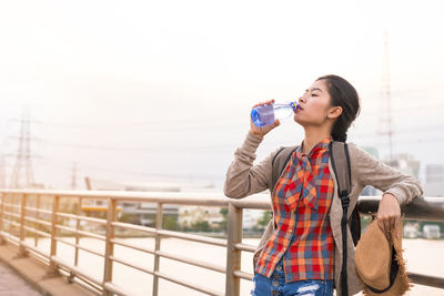 Young woman drinking water while standing against railing and sky