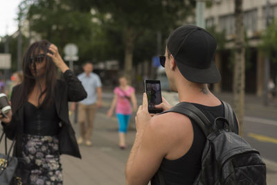 Man photographing friend with smart phone standing on footpath in city