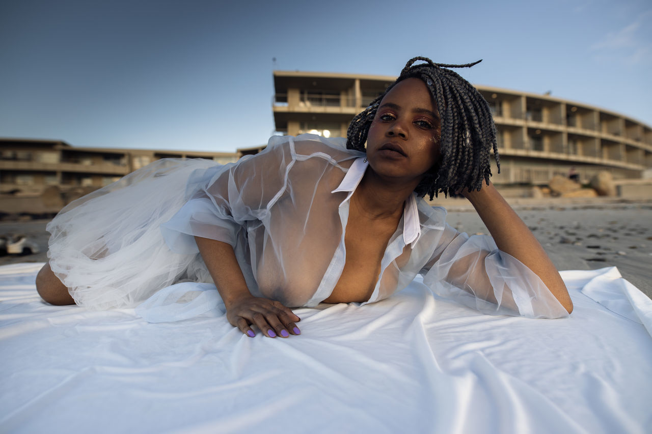 PORTRAIT OF BEAUTIFUL YOUNG WOMAN SITTING ON BED AGAINST SKY