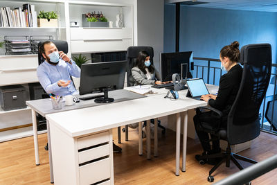 Serious young man in medical mask discussing business issue by phone while female colleagues working with computers in contemporary coworking office