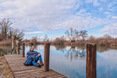 Mature man looking at view while relaxing on jetty over lake against cloudy sky