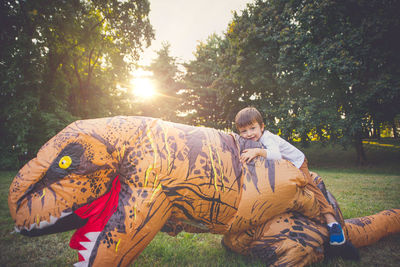 Portrait of boy sitting on person wearing dinosaur costume in park