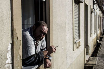Portrait of mature man smoking cigarette while leaning on window sill