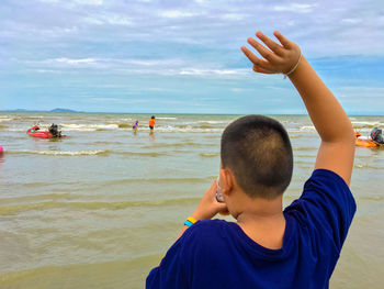 Rear view of man standing with arms raised on shore against cloudy sky