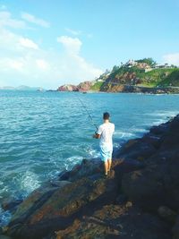 Rear view of man standing on rock at sea shore against sky