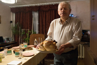 Casual gray haired male pensioner holding plate with roasted chicken while standing next to table arranged for family christmas dinner at home