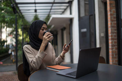 Young woman with laptop on table using smart phone while sitting in cafe