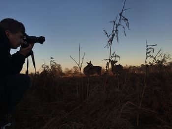 Man photographing deer on field against sky during sunset