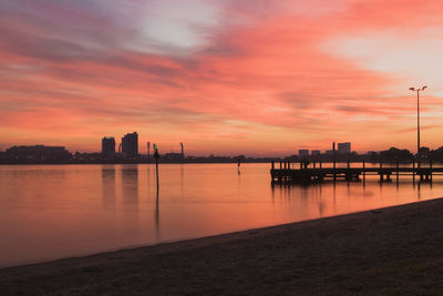 Silhouette buildings by sea against orange sky