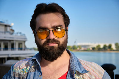 Portrait of young adult bearded caucasian man wearing orange sunglasses opposite river