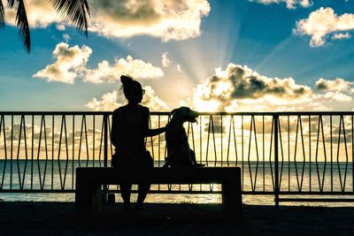 Silhouette woman with dog sitting by railing against sea during sunset