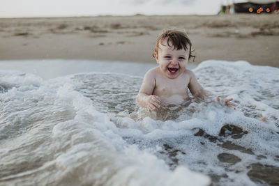 Male toddler laughing while sitting in water at beach during sunset