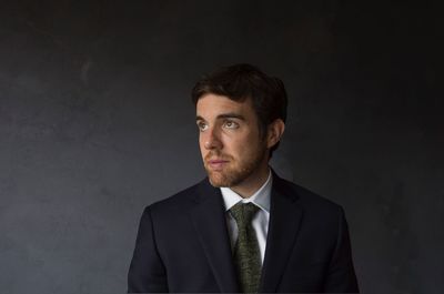 Thoughtful young businessman standing in suit by black wall