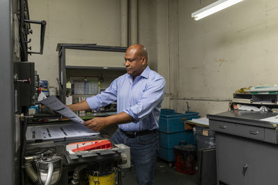 Male employee examining paper by print machine in shop