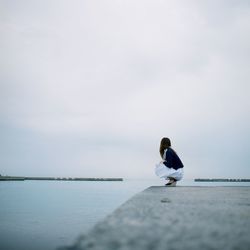 Side view of woman crouching on pier over lake against sky