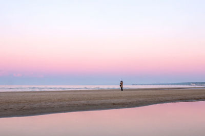 A photographer stands on the wide deserted sea beach surrounded by pink evening light