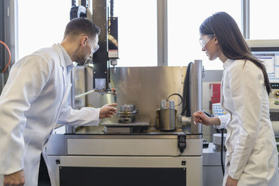 Colleagues wearing lab coats and safety goggles looking at machine in modern factory