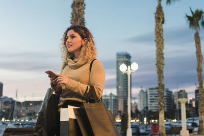 Side view of happy young woman looking away with shopping bags laughing while texting on mobile phone with modern city in twilight on background