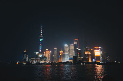 Illuminated buildings by river against clear sky at night