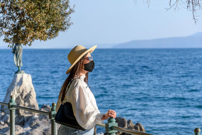 Portrait of a young woman wearing black protective mask, standing on shore of sea.