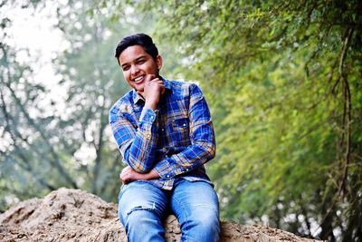 Portrait of smiling young man sitting on rock in forest