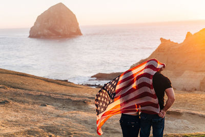 Rear view of man and woman with american flag standing at beach against sky