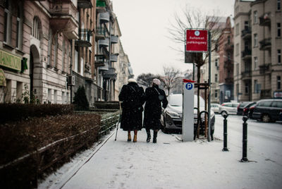 Rear view of people walking on snow covered sidewalk in city against sky