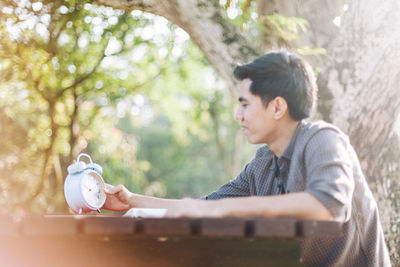 Young man looking at alarm clock while sitting at table in park
