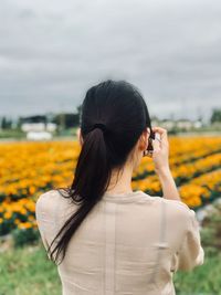 Rear view of young woman photographing with mobile phone while standing on field against cloudy sky