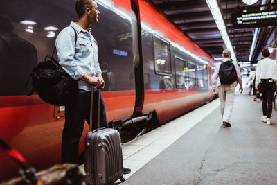 Male tourist with luggage looking away while standing against train on railroad station platform