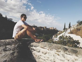 Portrait of man sitting on retaining wall against sky