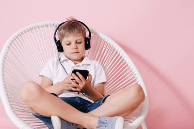 Boy listening music while using phone on chair against colored background