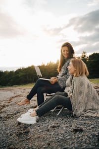 Smiling woman holding laptop while sitting with friend at beach during cold weather